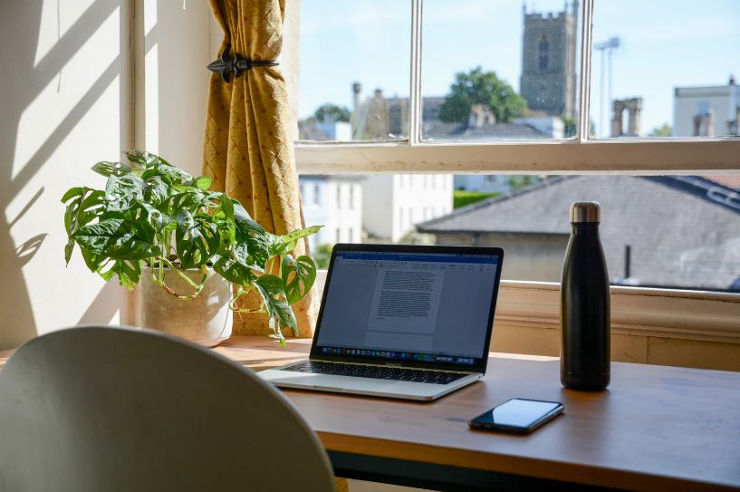 A laptop and a bottle on a desk in front of a window overlooking some buildings as well as a possible castle or fort in the distance.